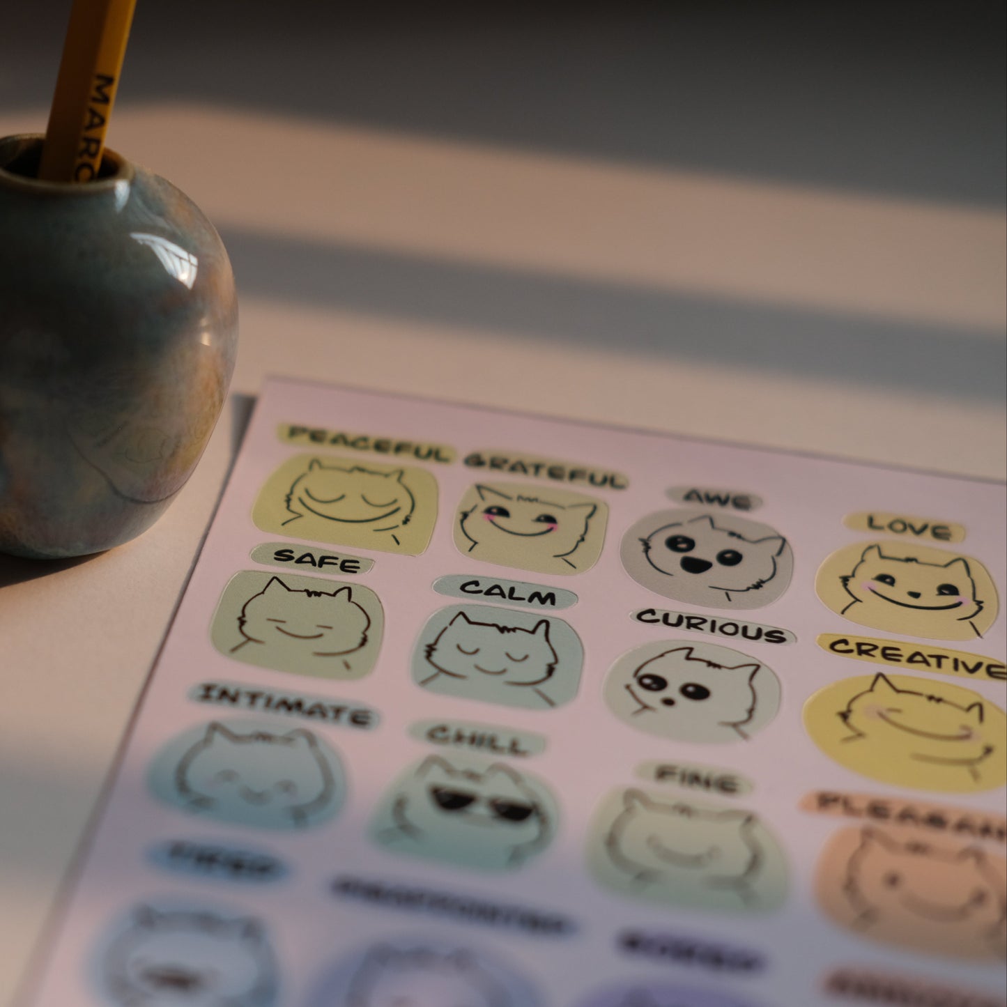 Small figurine next to a pen holder with a sheet of stickers featuring cat illustrations on a desk.
