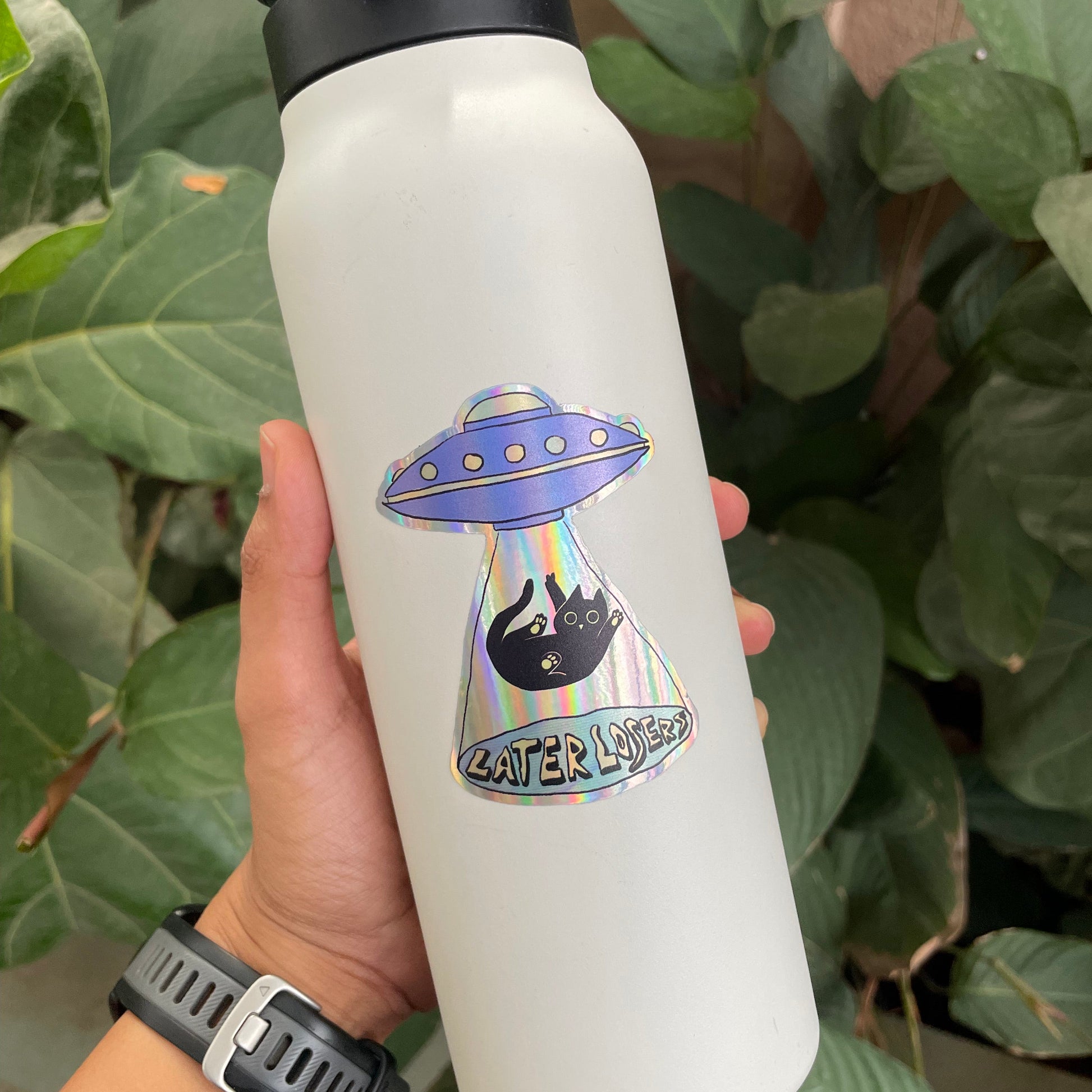 White water bottle with a UFO and cat design held by a person against a green leafy background