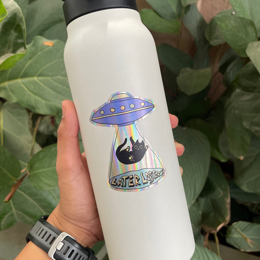 White water bottle with a UFO and cat design held by a person against a green leafy background