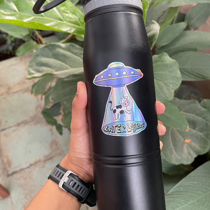 Black water bottle with a colorful UFO & Cow holographic sticker held by a person against a green leafy background