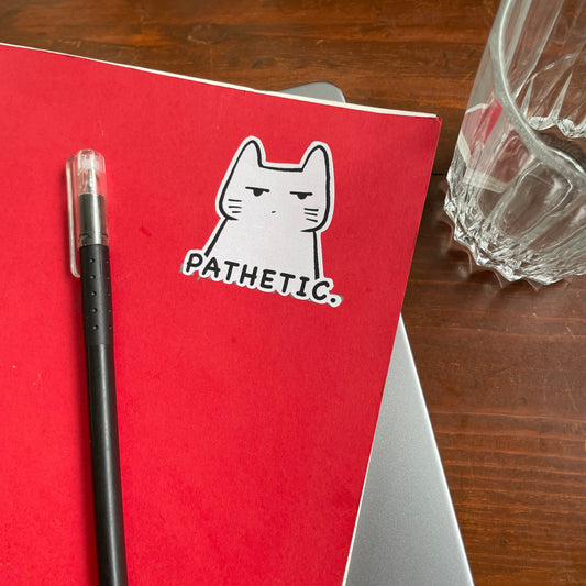 Red notebook with a cat sticker labeled 'Pathetic' on a wooden surface with a glass and pen.
