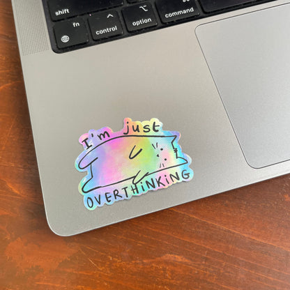 Holographic cat sticker on a laptop with text 'I'm just overthinking' on a wooden surface