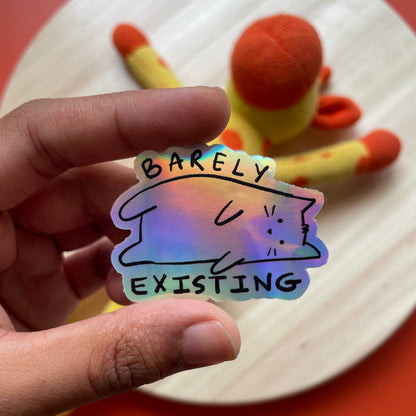 Holographic sticker with a cat design and text held by a hand against a colorful background