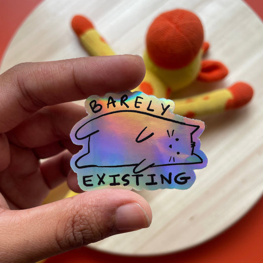 Holographic sticker with a cat design and text held by a hand against a colorful background