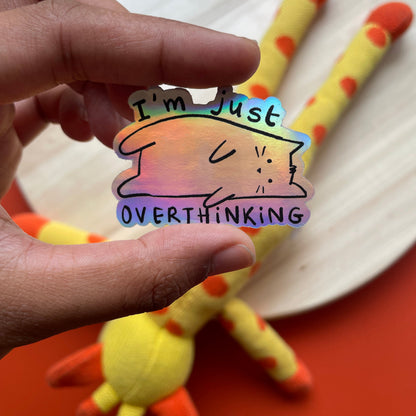 Hand holding a colorful sticker with a cat illustration and text, with a plush toy in the background.