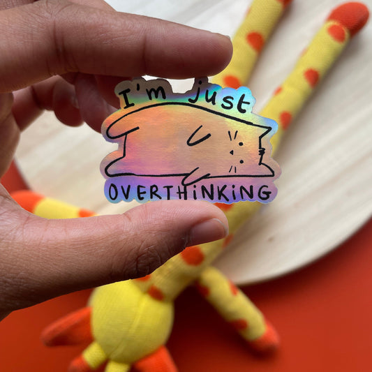 Hand holding a colorful sticker with a cat illustration and text, with a plush toy in the background.