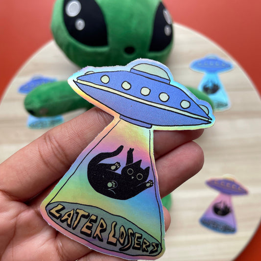 Holographic sticker with a cat and UFO design held by a hand, with a colorful alien toy in the background.