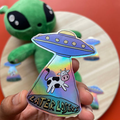 Holographic sticker with a cow and UFO design held by a hand, with a red background.