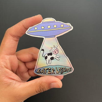 Hand holding a colorful UFO-shaped sticker with a cow design and text on a black background