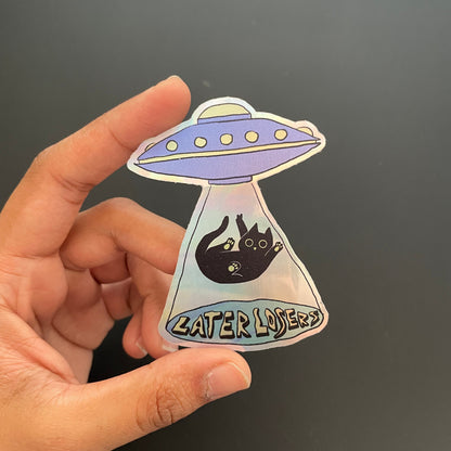 Hand holding a UFO-shaped sticker with cat and text on a black background