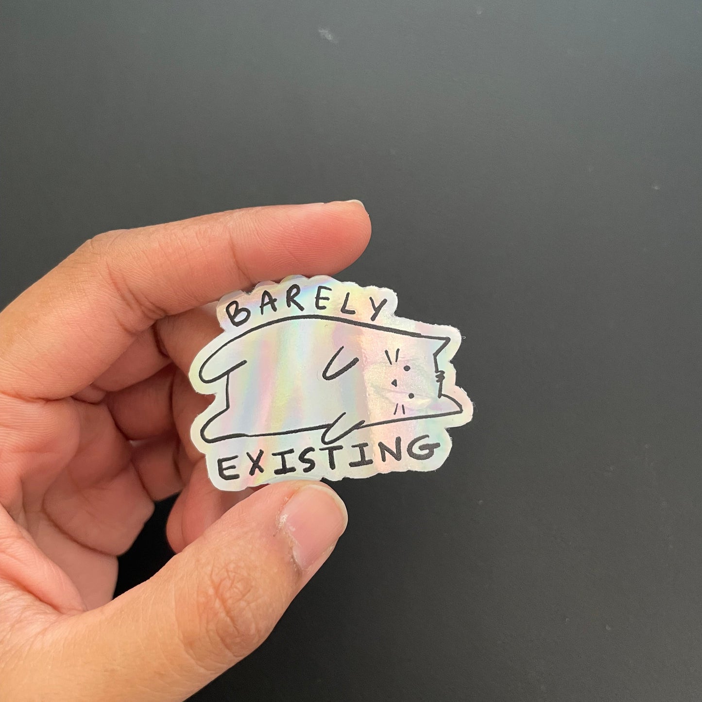 Hand holding a holographic sticker with a cat design and text on a black background
