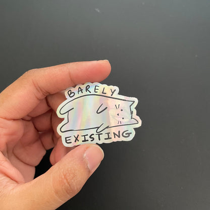 Hand holding a holographic sticker with a cat design and text on a black background