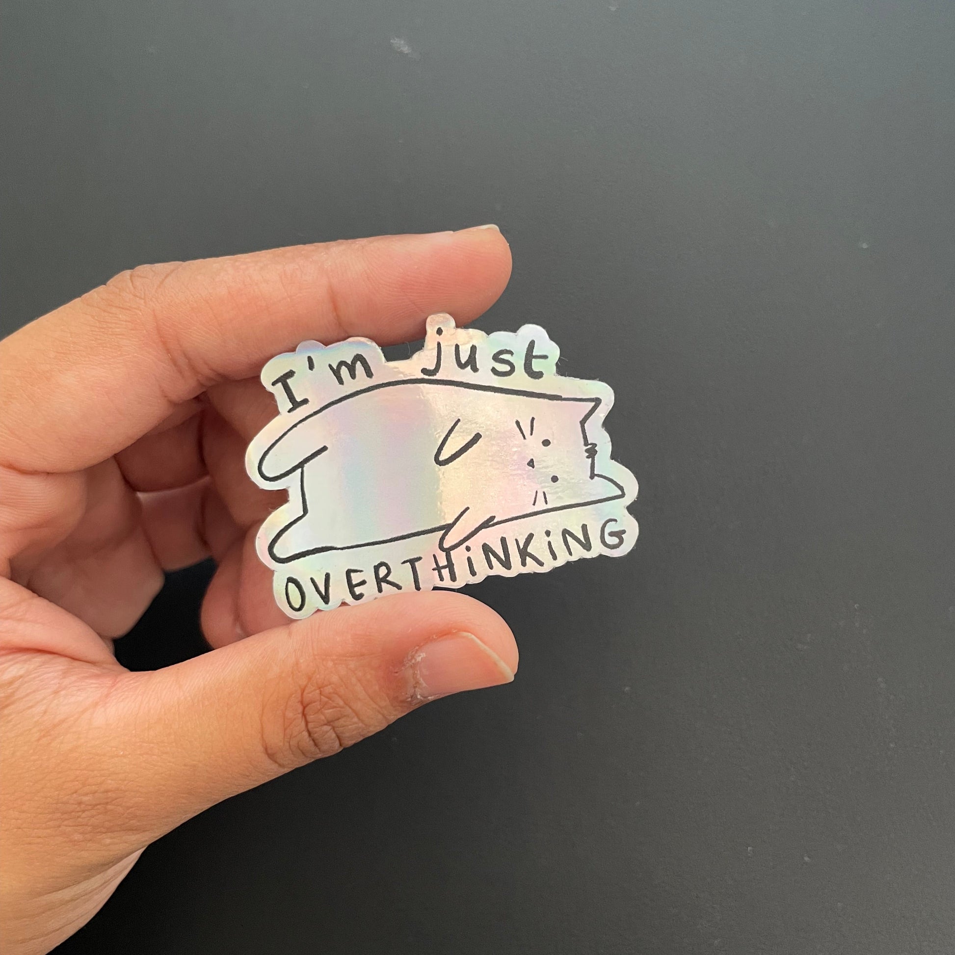Hand holding a holographic sticker with a cat design and text on a black background