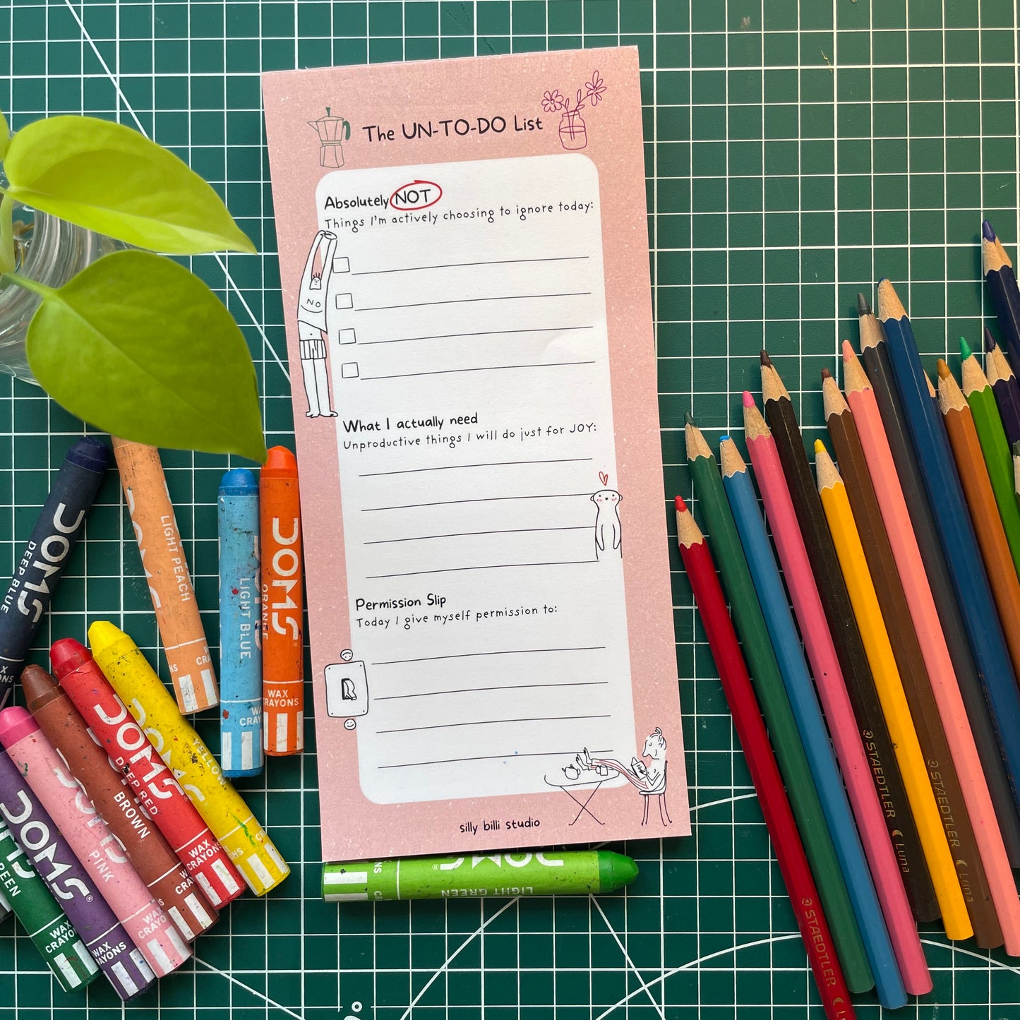Pink notepad titled 'The UN-TO-DO List' with colored pencils and markers on a green grid background