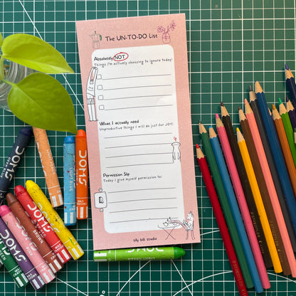 Pink notepad titled 'The UN-TO-DO List' with colored pencils and markers on a green grid background