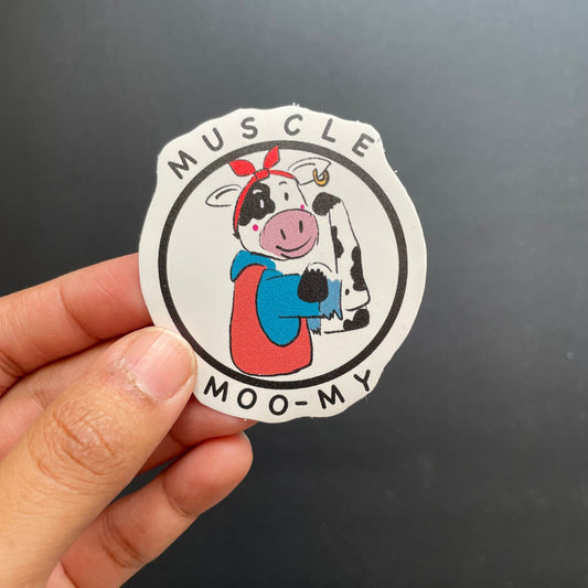 Sticker of a cow character with text 'Muscle Moo-My' held by a hand against a dark background